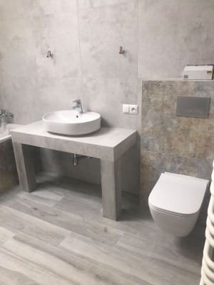 Wykończona łazienka z okrągłą umywalką na betonowym blacie, podwieszaną toaletą i płytkami imitującymi beton na ścianach i podłodze.