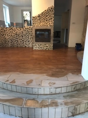 Wnętrze z drewnianą podłogą w trakcie renowacji, widoczne schody wyłożone jasnymi płytkami ceramicznymi i kominek obłożony dekoracyjnymi kamieniami.