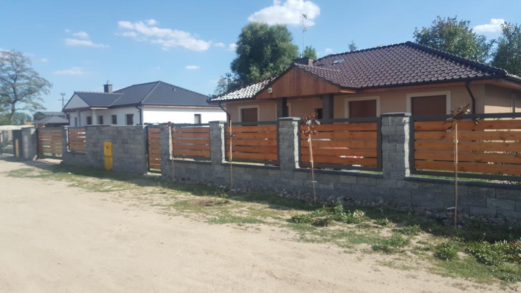 Nowoczesne ogrodzenie z poziomych desek drewnianych i słupków z szarej kostki, oddzielające posesje z domami jednorodzinnymi.