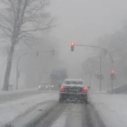 Samochód osobowy jadący po ośnieżonej drodze podczas intensywnych opadów śniegu, widoczne sygnalizacja świetlna i inne pojazdy w oddali.