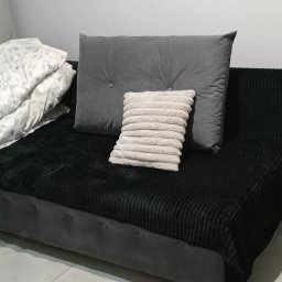 PRESTIGE EDYTA LANGHOLTZ - Ustawione poduszki i kołdra na szarym tapczanie z czarnym, prążkowanym pledem. Minimalistyczne wnętrze z jasną ścianą w tle.