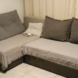 PRESTIGE EDYTA LANGHOLTZ - Szary narożnik z poduszkami i narzutą z frędzlami w jasnym wnętrzu. Widoczne detale: biała poduszka dekoracyjna i okulary leżące na siedzisku.