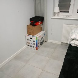 PRESTIGE EDYTA LANGHOLTZ - Pokój z ułożonymi kartonami i torbą obok ściany, grzejnika i łóżka. Na podłodze szare płytki. Widoczne okno z lampką.
