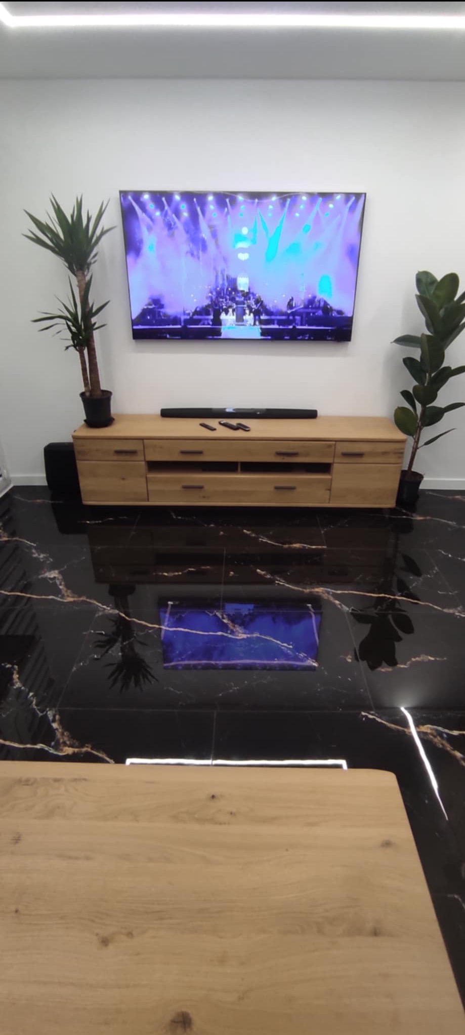 Salon z czarną, marmurkową podłogą, drewnianą szafką RTV, telewizorem i roślinami doniczkowymi. Nowoczesne wnętrze z odbiciem w podłodze.