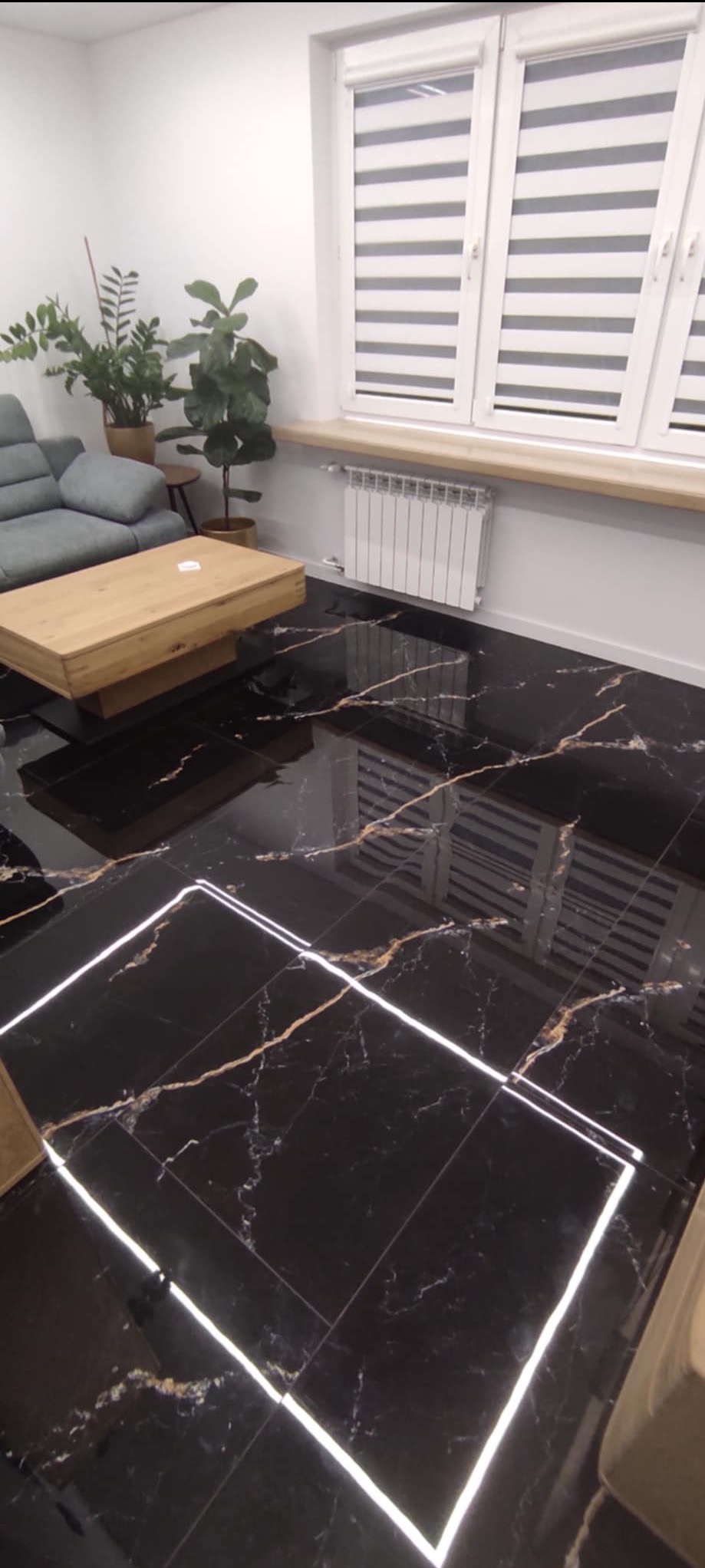 Salon w Płocku z czarną, marmurkową podłogą z podświetleniem LED. Widoczny drewniany stolik kawowy, szara sofa i rośliny doniczkowe przy oknie z żaluzjami.