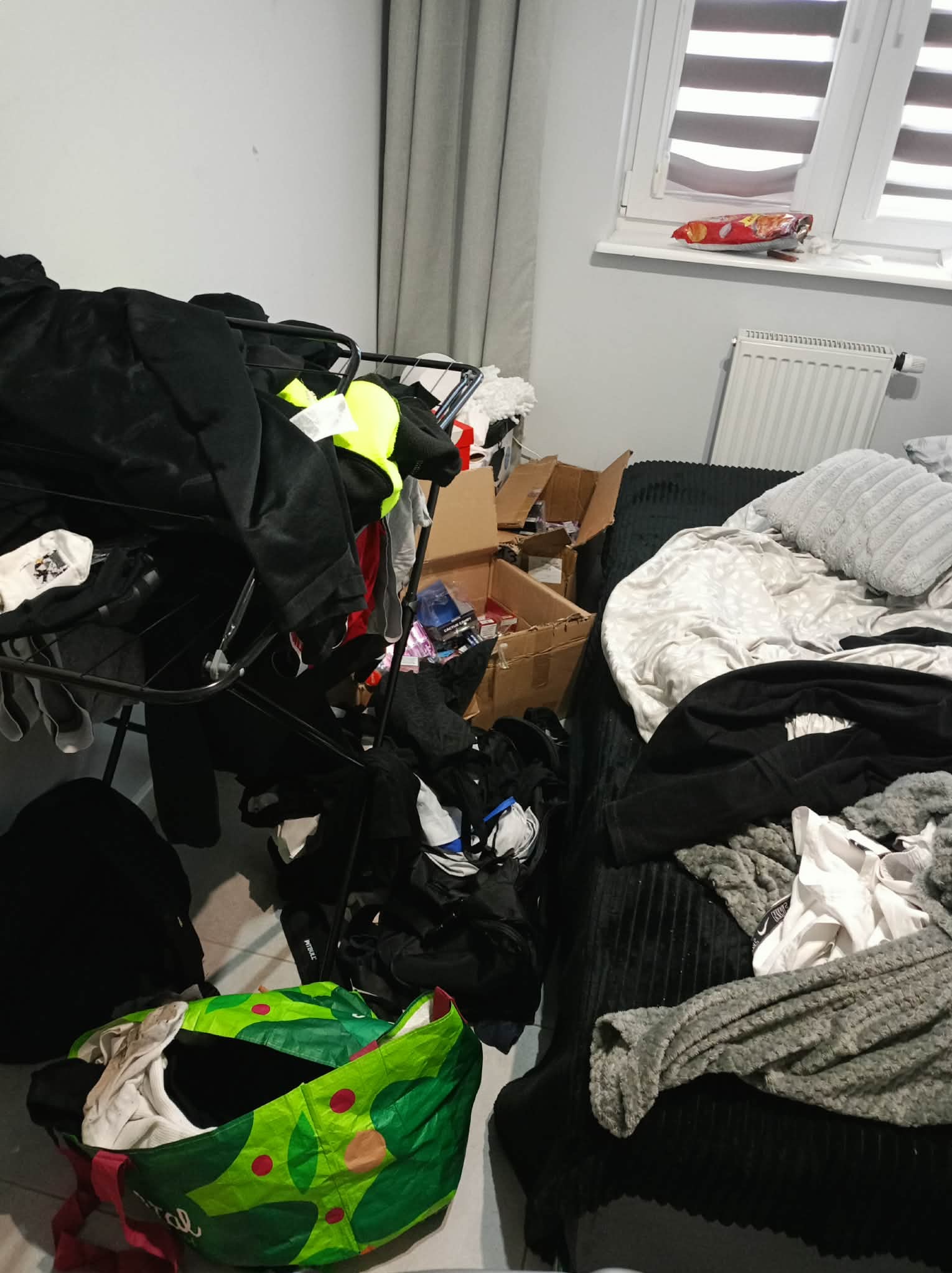 Zagracony pokój: ubrania na suszarce, kartony, torba na podłodze, niepościelone łóżko. Bałagan w pokoju z oknem i grzejnikiem.