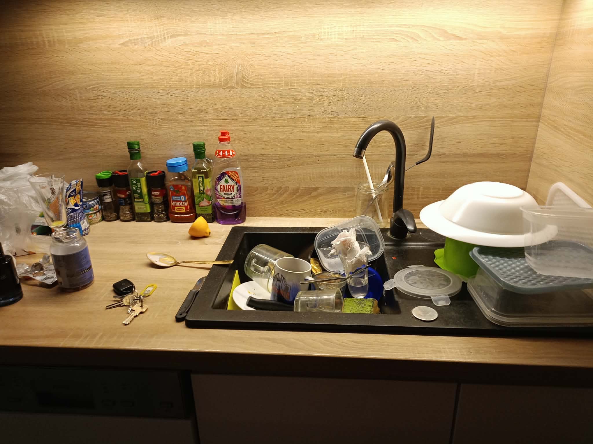 Zlew pełen brudnych naczyń, resztek jedzenia i środków czystości na blacie kuchennym z drewnianym wykończeniem. Widoczne przyprawy, klucze i niedomyte naczynia.