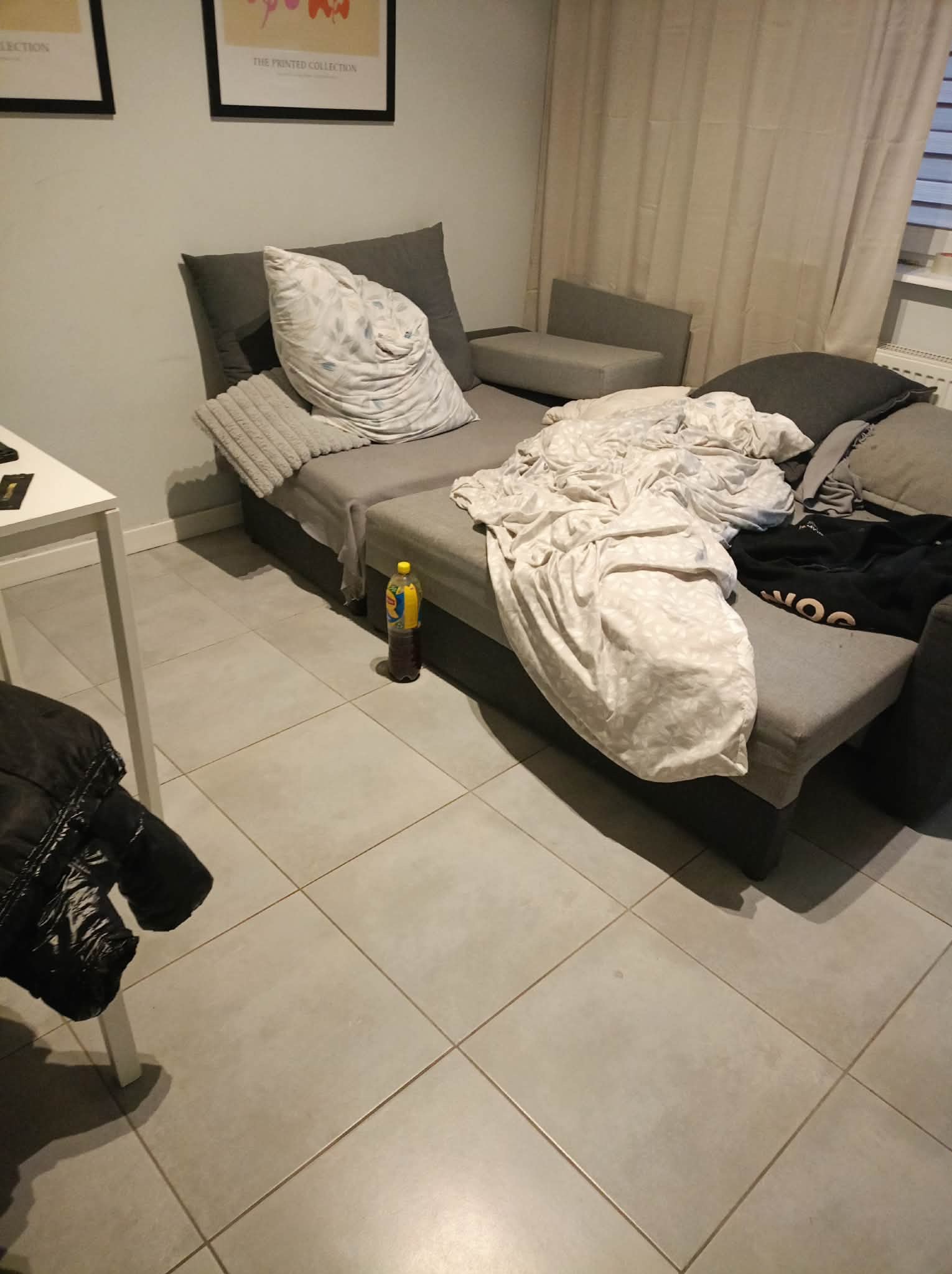 Niedbale posłane łóżko z butelką napoju na podłodze, w tle zasłony i plakaty na ścianie. Widok na wnętrze pokoju przed sprzątaniem.