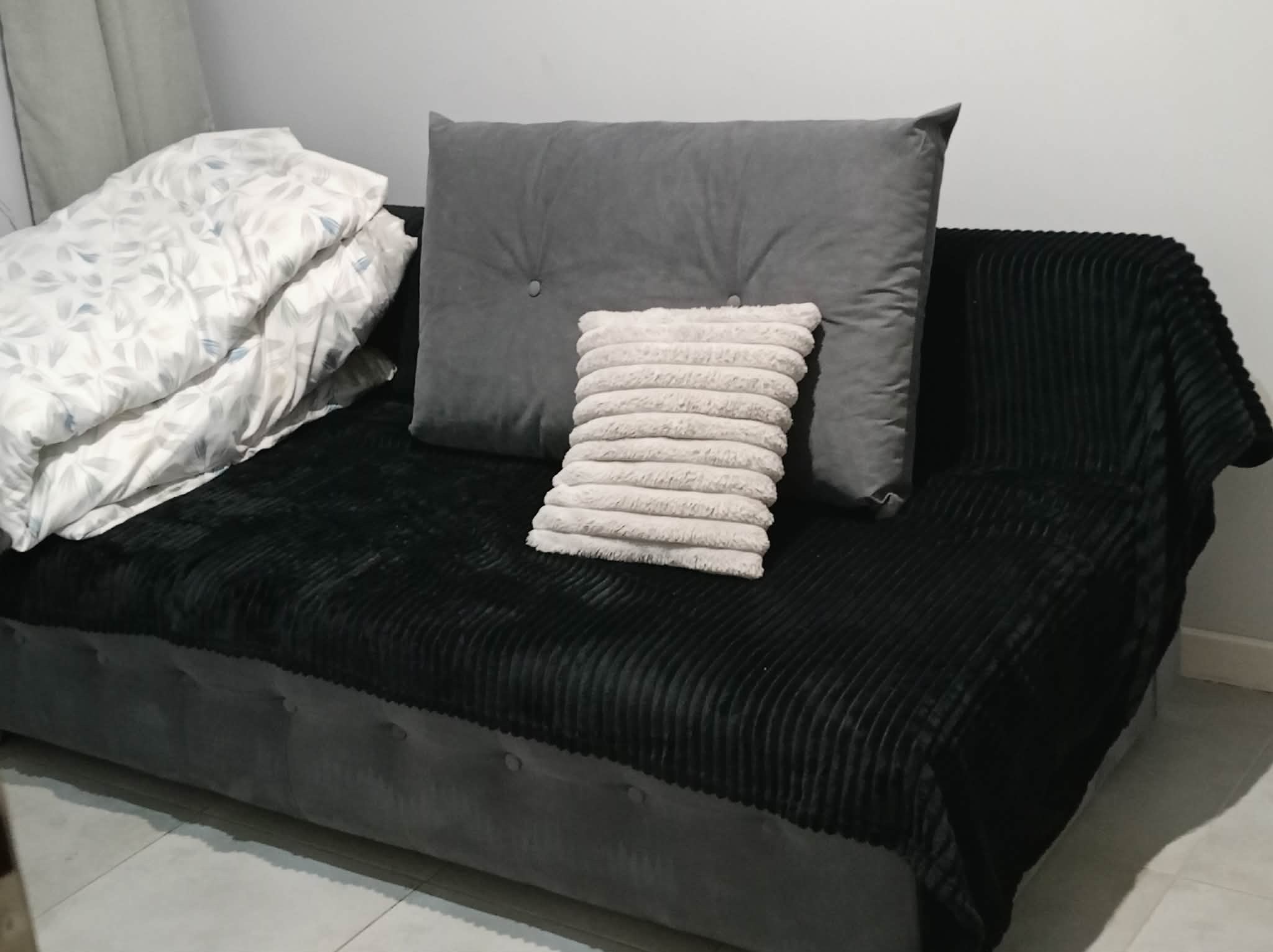 Ustawione poduszki i kołdra na szarym tapczanie z czarnym, prążkowanym pledem. Minimalistyczne wnętrze z jasną ścianą w tle.