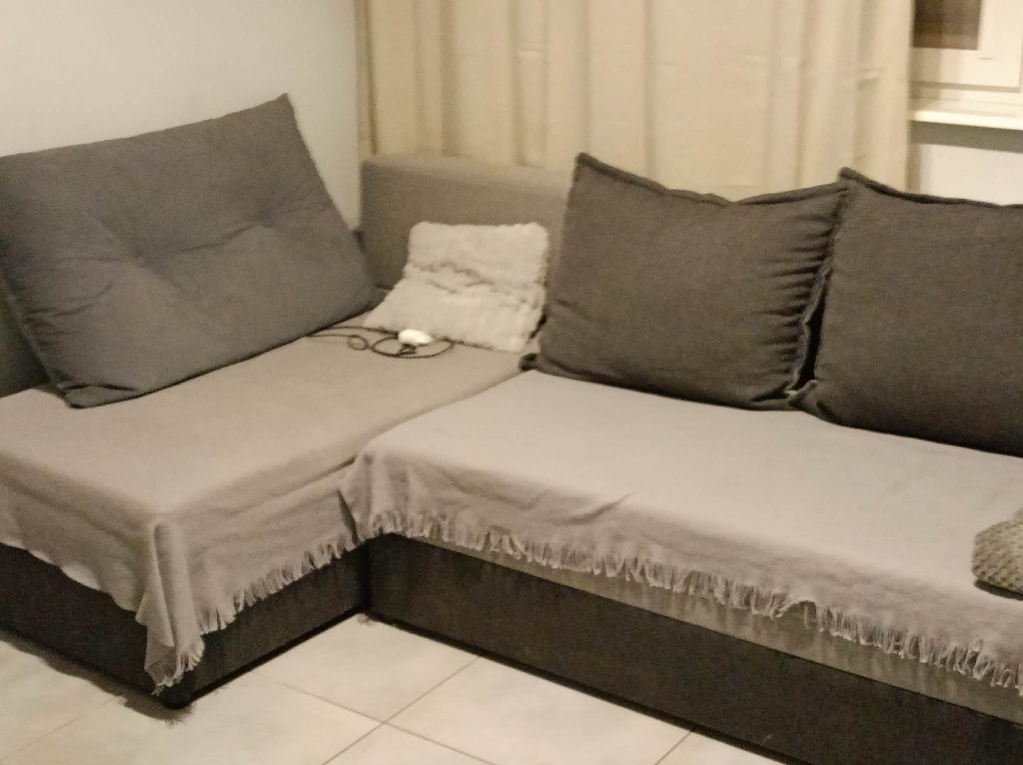 Szary narożnik z poduszkami i narzutą z frędzlami w jasnym wnętrzu. Widoczne detale: biała poduszka dekoracyjna i okulary leżące na siedzisku.