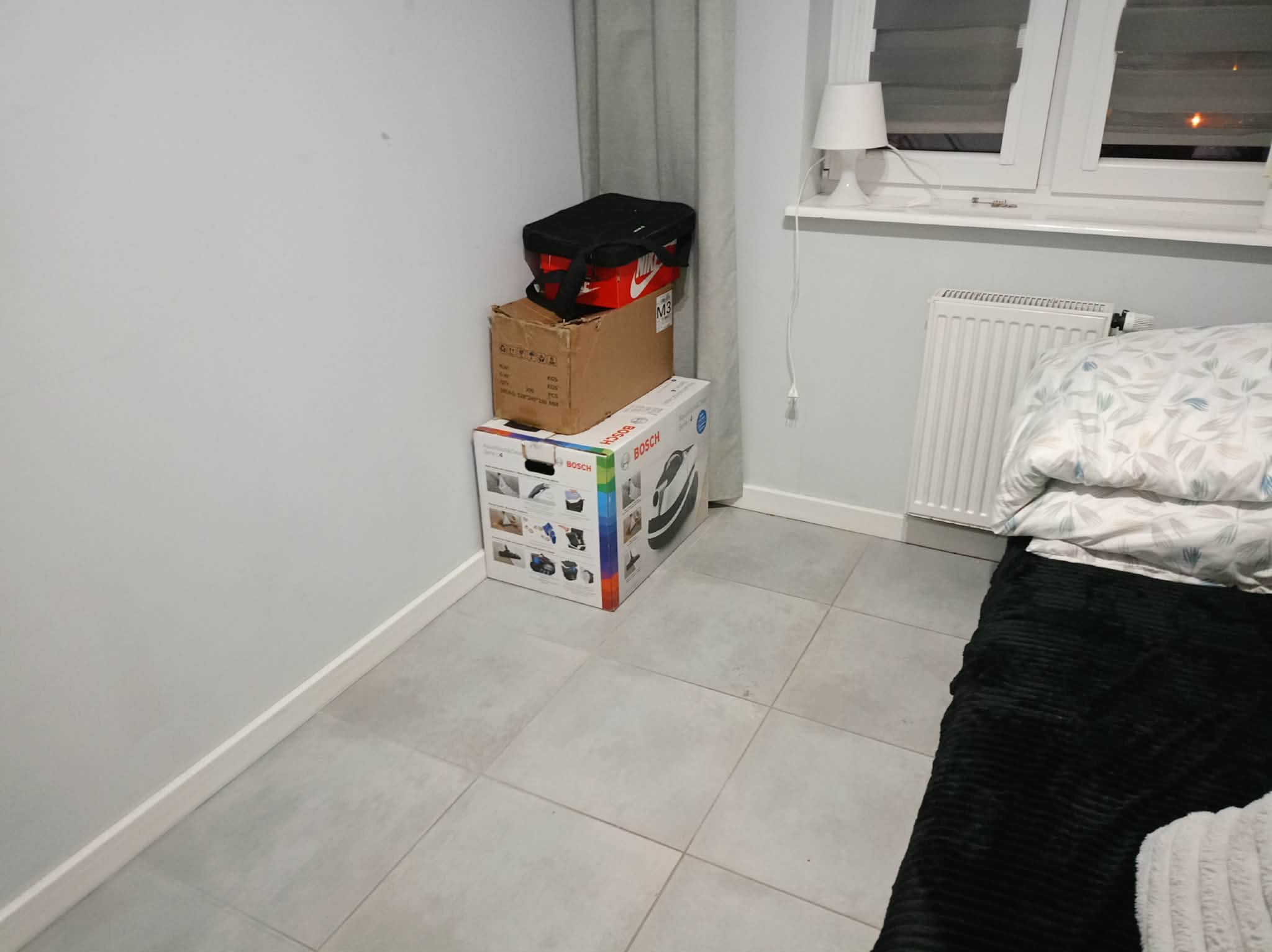 Pokój z ułożonymi kartonami i torbą obok ściany, grzejnika i łóżka. Na podłodze szare płytki. Widoczne okno z lampką.