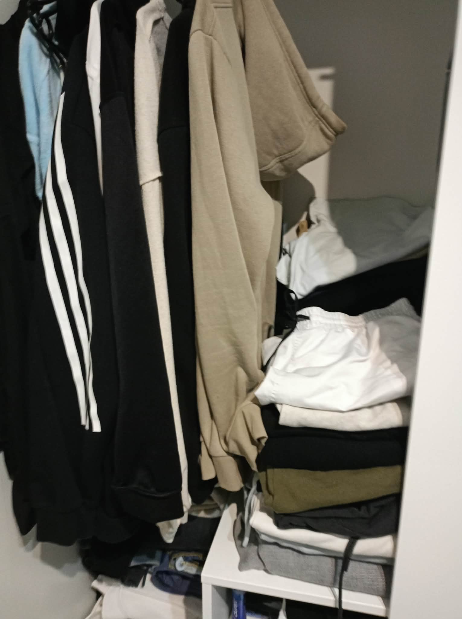 Uporządkowana garderoba z wiszącymi bluzami i poskładanymi ubraniami na półkach. Widoczne ubrania sportowe i codzienne w stonowanych kolorach.