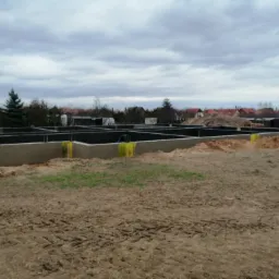 Fundamenty domu jednorodzinnego w trakcie budowy, z widocznymi żółtymi zabezpieczeniami na izolacji przeciwwilgociowej, ziemia wokół wykopu, pochmurne niebo w tle.