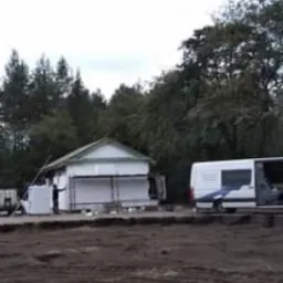 Mały, parterowy dom w trakcie ocieplania elewacji, widoczny pracownik przy ścianie, teren wokół rozkopany, w tle las i samochód dostawczy.