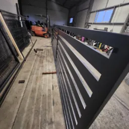 Czarna, nowoczesna brama z poziomymi szczeblami w warsztacie, w tle wózek widłowy i elementy ogrodzeń.