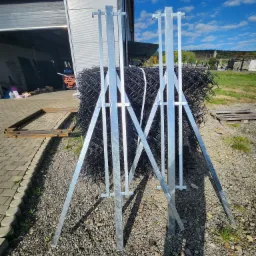 Zwój czarnej siatki ogrodzeniowej podparty na dwóch metalowych stojakach, na żwirowym podłożu, w tle budynek z otwartą bramą i błękitne niebo.