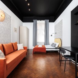 UPSTAIRS MANAGEMENT SPÓŁKA Z OGRANICZONĄ ODPOWIEDZIALNOŚCIĄ - Jasne wnętrze mieszkania z parkietem w jodełkę, pomarańczową sofą, łóżkiem i czarnym biurkiem. Ściany w biało-szare wzory, czarny sufit z oświetleniem punktowym.