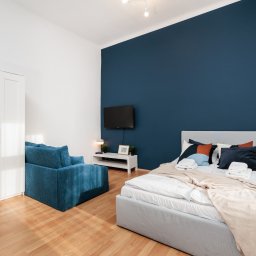 UPSTAIRS MANAGEMENT SPÓŁKA Z OGRANICZONĄ ODPOWIEDZIALNOŚCIĄ - Pokój z łóżkiem, sofą i szafą w odcieniach bieli i błękitu. Ściana za łóżkiem pomalowana na granatowo. Na ścianie wisi telewizor. Parkiet na podłodze.
