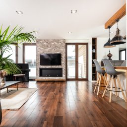 UPSTAIRS MANAGEMENT SPÓŁKA Z OGRANICZONĄ ODPOWIEDZIALNOŚCIĄ - Nowoczesny salon z parkietem, ceglaną ścianą z TV i kominkiem, pianinem, rośliną doniczkową i barkiem z krzesłami. Jasne, przestronne wnętrze.