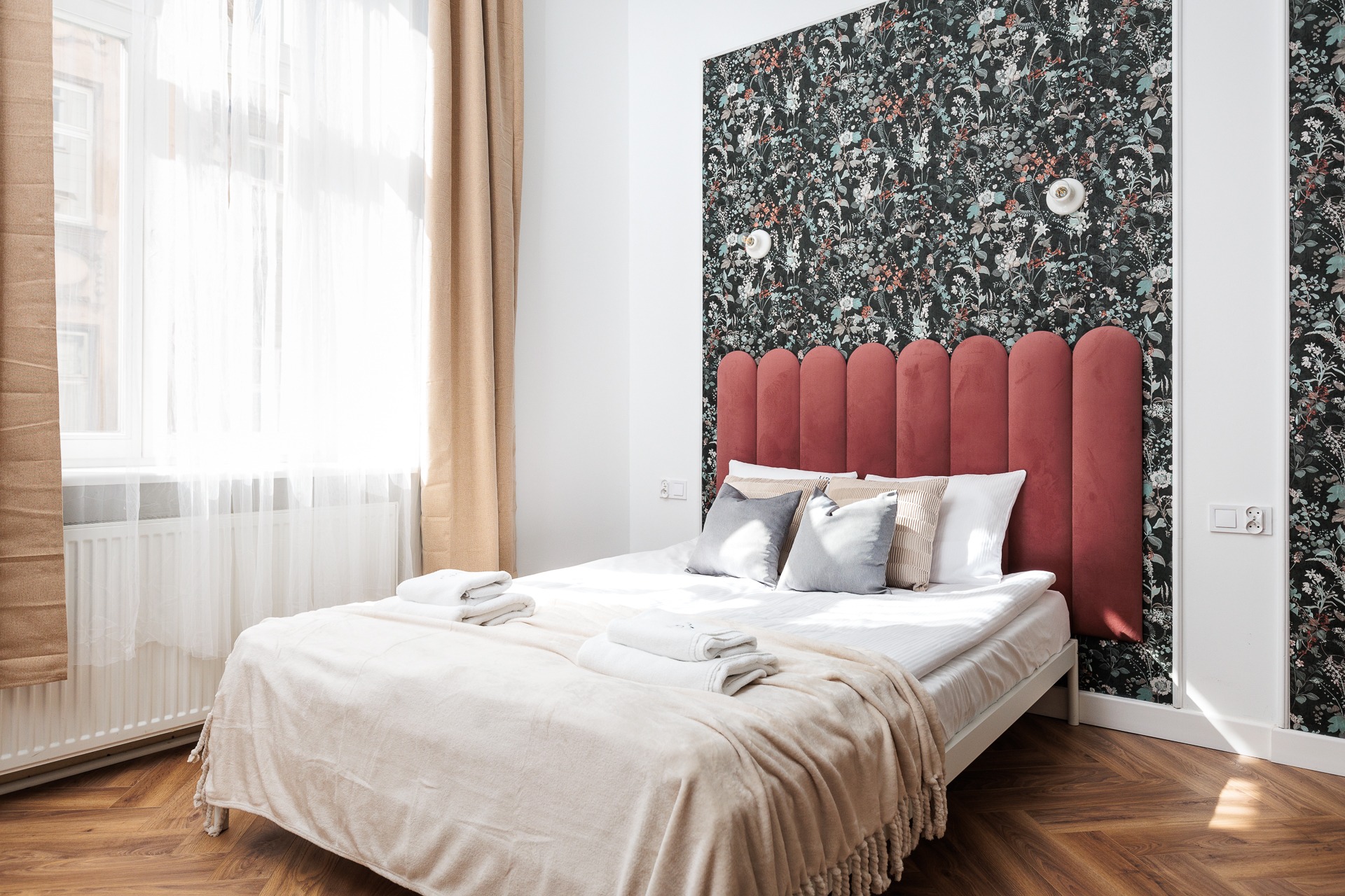 Jasna sypialnia z wzorzystą tapetą, czerwonym zagłówkiem i beżowymi zasłonami. Na łóżku leżą ręczniki i poduszki. Parkiet odbija światło słoneczne.
