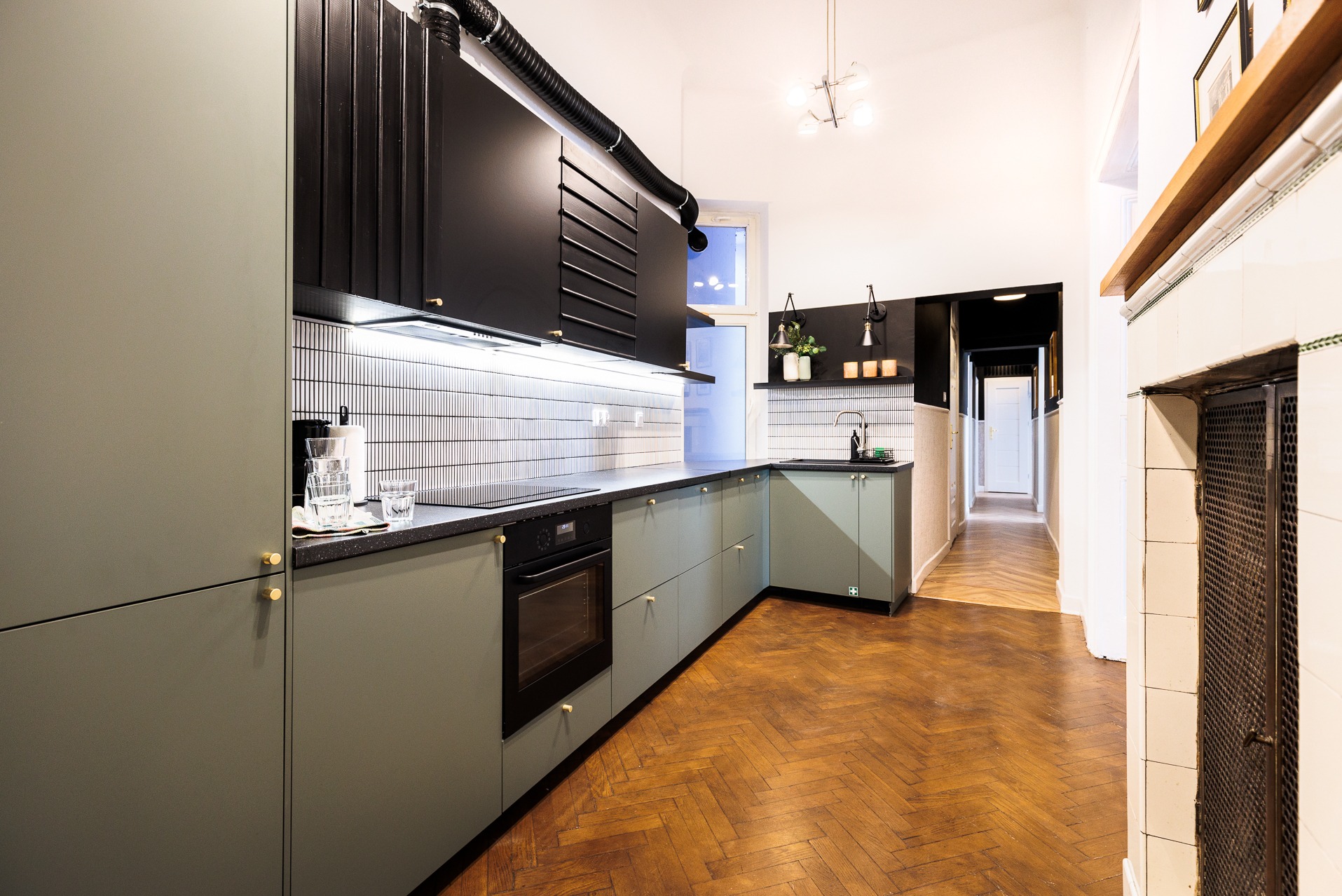 Nowoczesna kuchnia w odcieniach zieleni i czerni, z parkietem w jodełkę. Widoczny piekarnik, szafki, czarne górne zabudowy i fragment korytarza.
