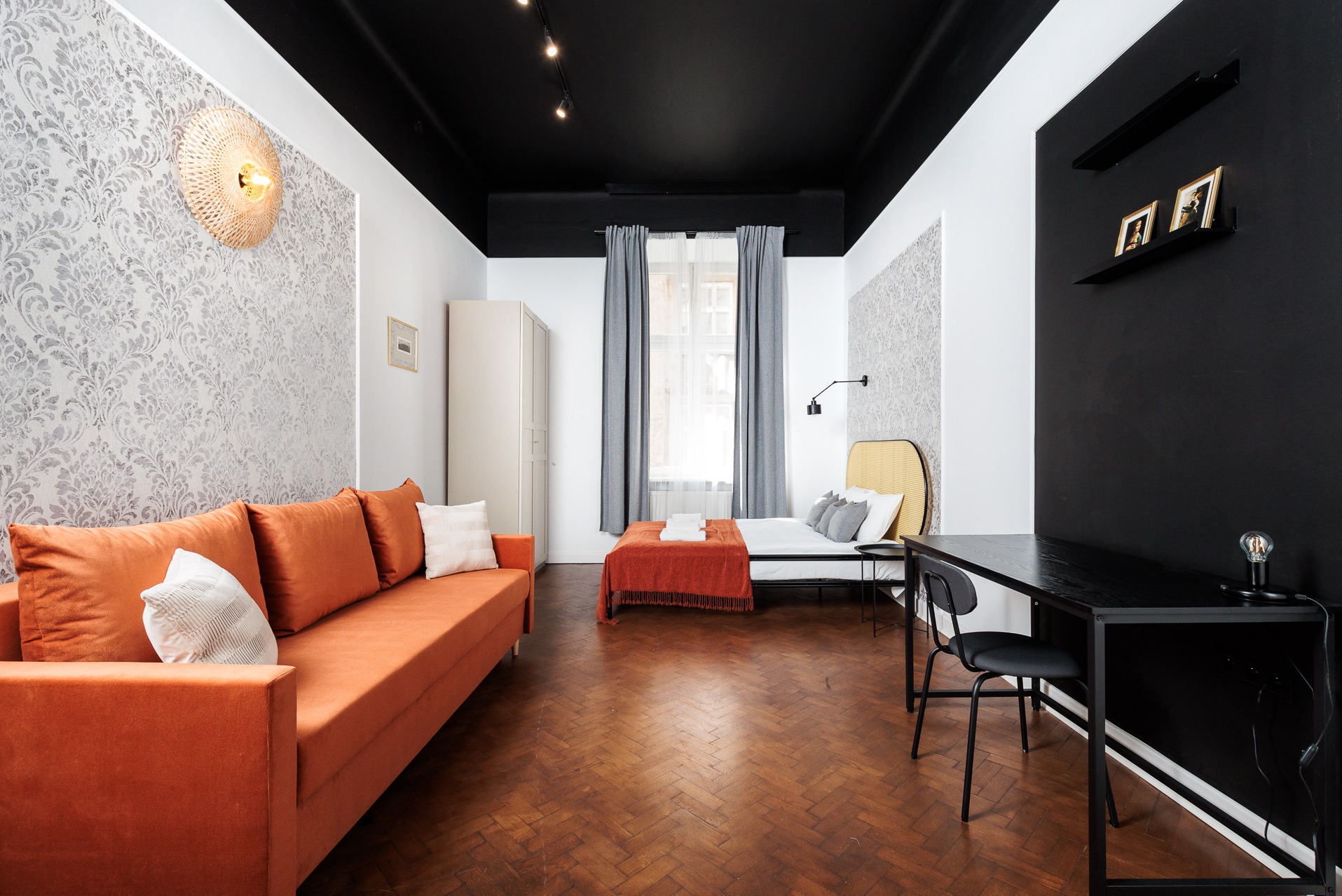 Jasne wnętrze mieszkania z parkietem w jodełkę, pomarańczową sofą, łóżkiem i czarnym biurkiem. Ściany w biało-szare wzory, czarny sufit z oświetleniem punktowym.