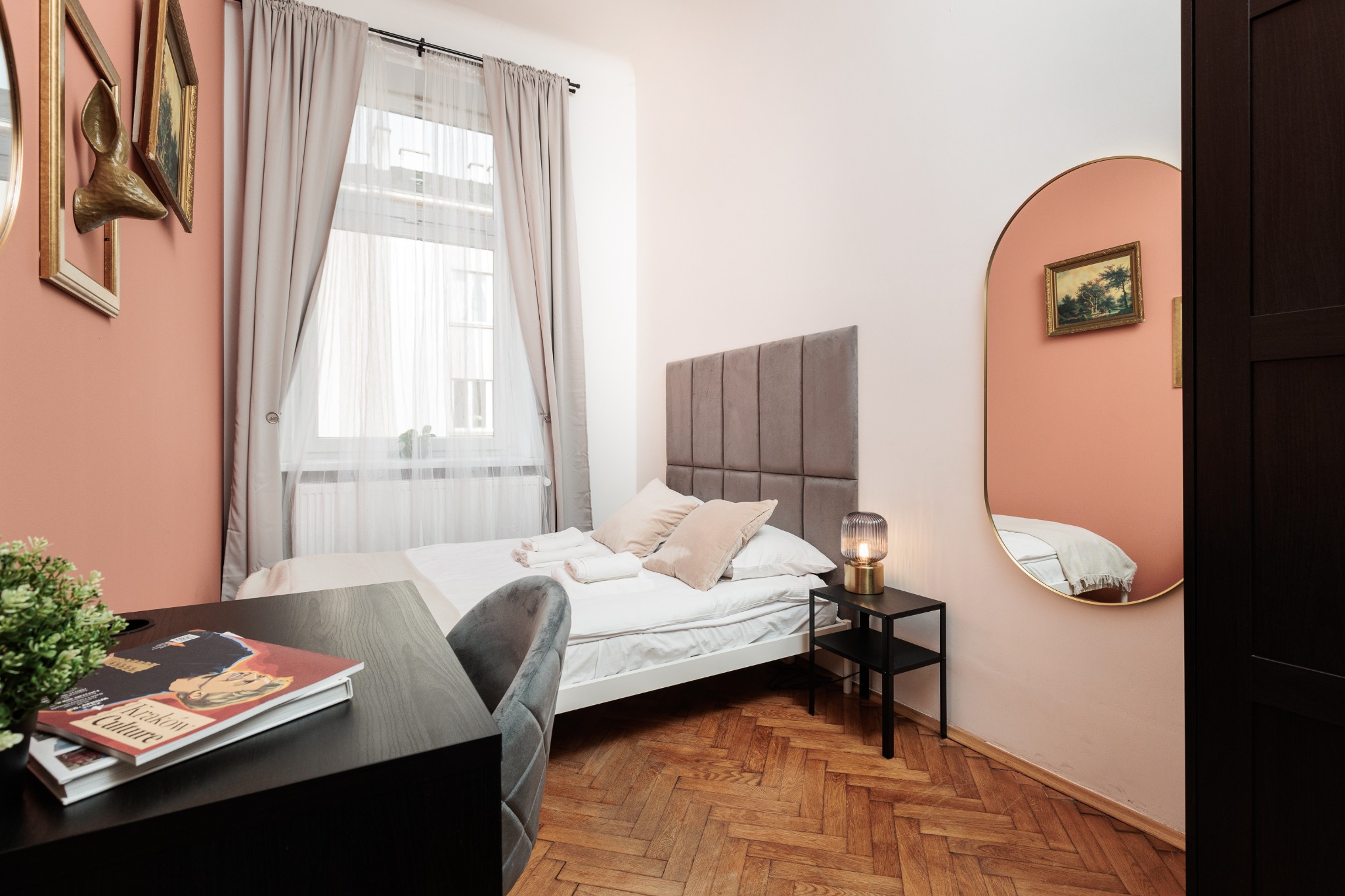 Przytulna sypialnia w odcieniach różu i szarości z drewnianą podłogą. Na biurku magazyn 'Krakow Culture'. Lustro owalne odbija obraz na ścianie.