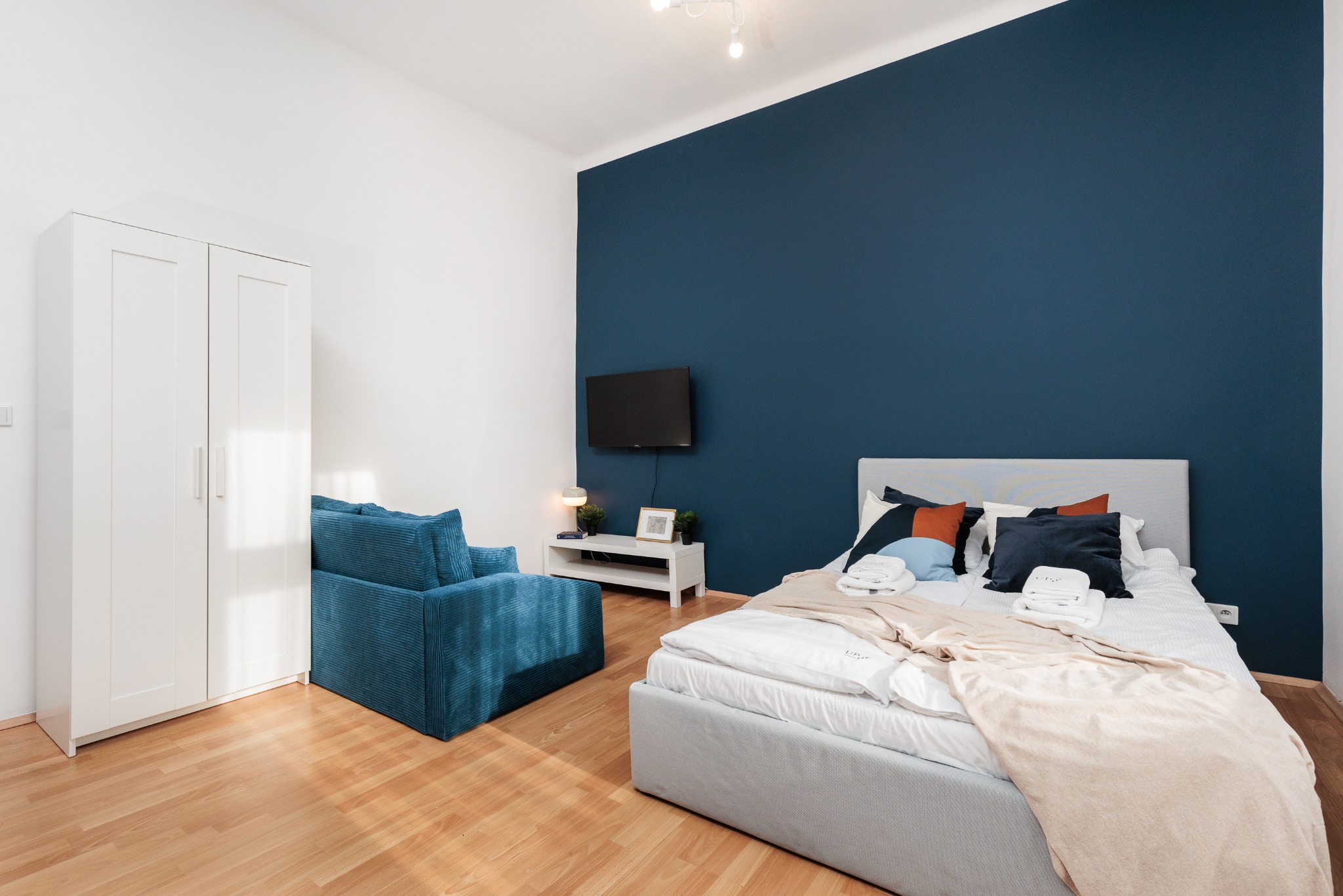 Pokój z łóżkiem, sofą i szafą w odcieniach bieli i błękitu. Ściana za łóżkiem pomalowana na granatowo. Na ścianie wisi telewizor. Parkiet na podłodze.