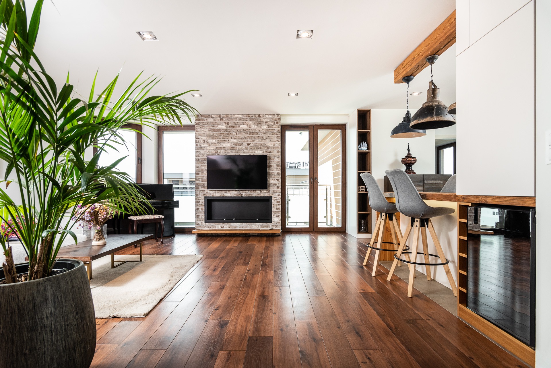 Nowoczesny salon z parkietem, ceglaną ścianą z TV i kominkiem, pianinem, rośliną doniczkową i barkiem z krzesłami. Jasne, przestronne wnętrze.