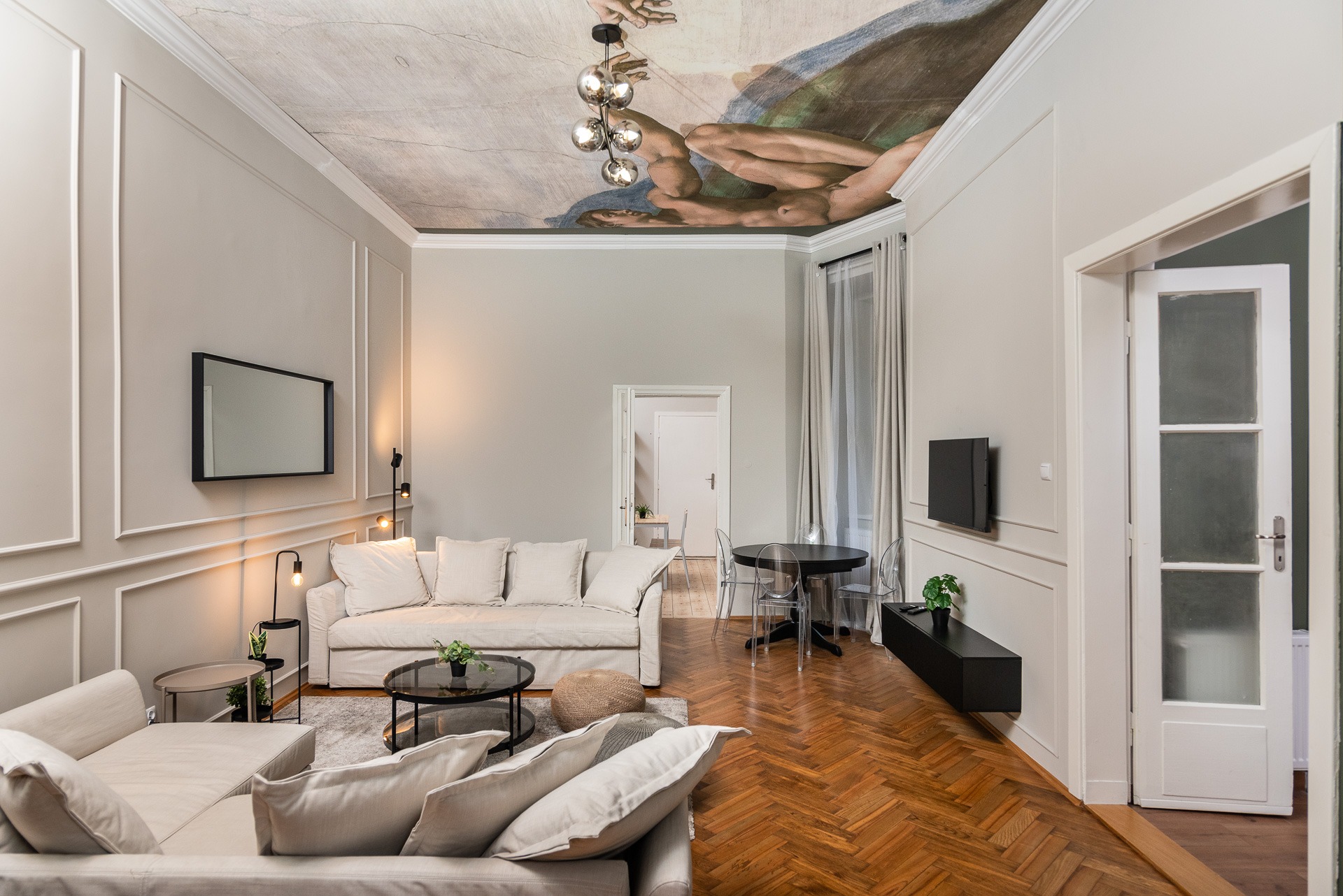 Jasny salon z parkietem w jodełkę, sofami i malowidłem na suficie w stylu Kaplicy Sykstyńskiej, oświetlony nowoczesną lampą.