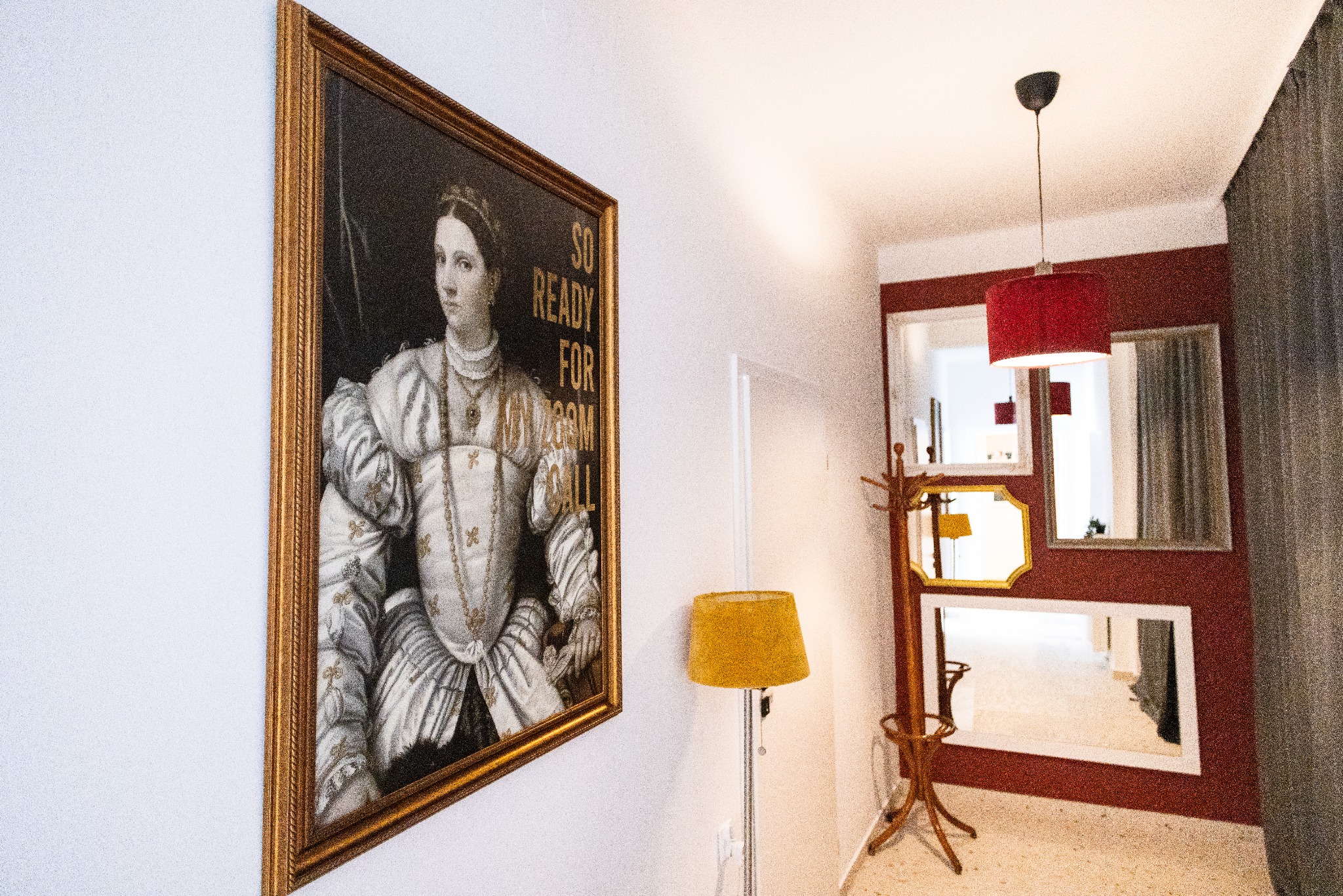 Korytarz z obrazem w złotej ramie przedstawiającym kobietę w renesansowym stroju z napisem 'SO READY FOR ROOM CALL', lampą i wieszakiem na ubrania.