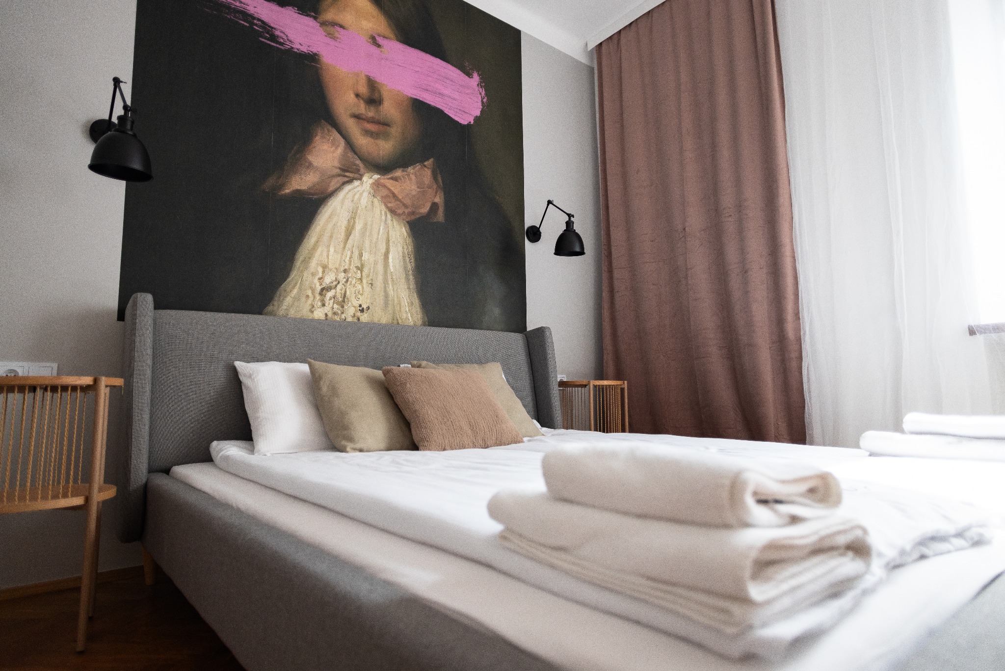 Pokój z łóżkiem, nad którym wisi obraz z różową plamą na twarzy postaci. Obok łóżka stoi krzesło, a w tle widać zasłony. Lampy ścienne po bokach obrazu.