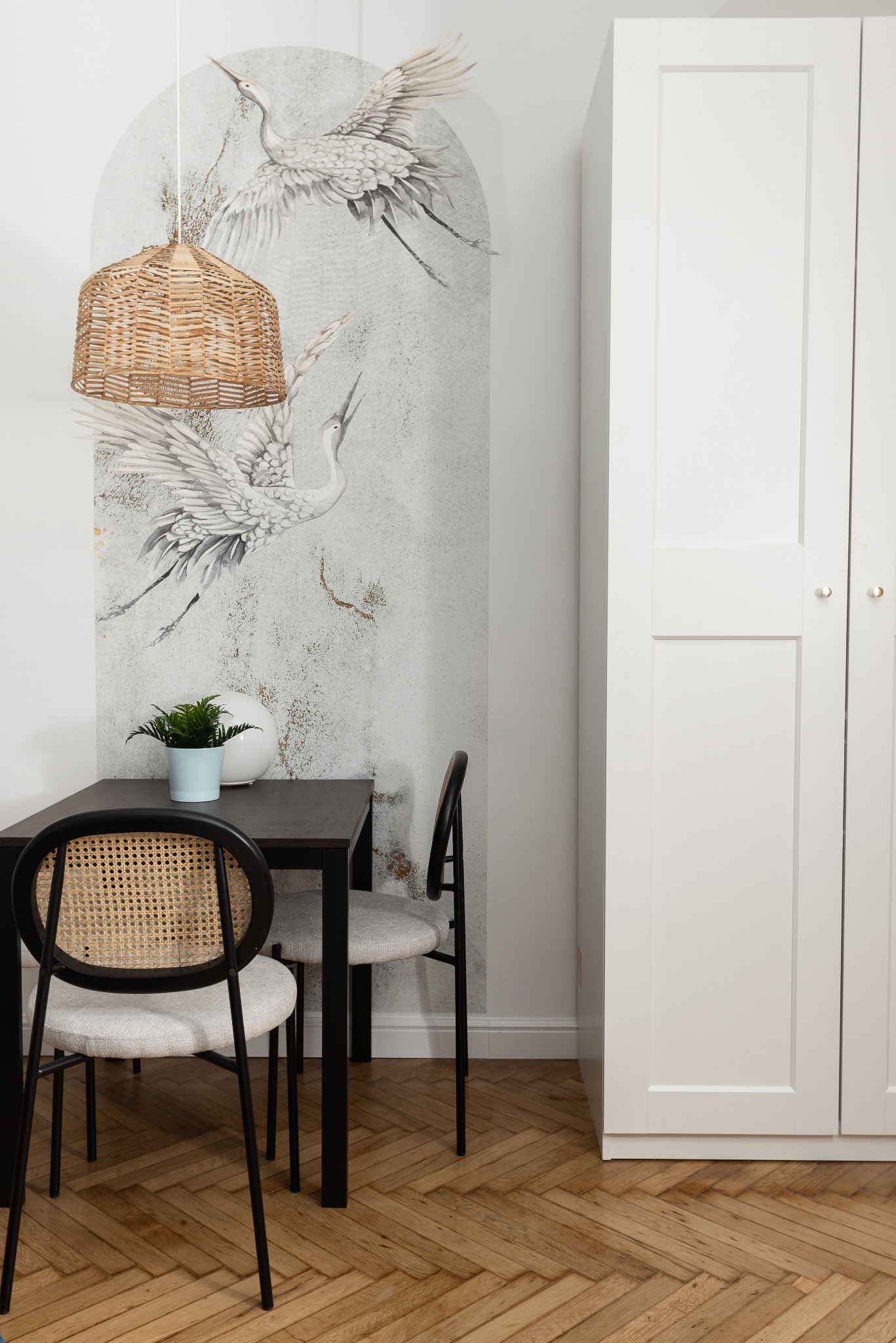 Stylowa aranżacja wnętrza: czarny stół z rattanowymi krzesłami, pleciona lampa, grafika z żurawiami na ścianie i parkiet w jodełkę. Minimalistyczny design.