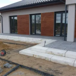 Nowoczesny dom z elewacją w kolorze beżowym i drewnopodobnymi panelami, widoczne schody wejściowe z jasnych bloków w trakcie budowy, kostka brukowa częściowo ułożona, w tle las.