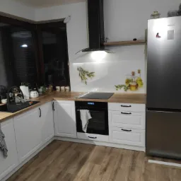 Nowoczesna kuchnia z białymi szafkami, drewnianym blatem i czarnym okapem, uzupełniona o szklany panel nad blatem z motywem oliwek i butelek oleju, a także o srebrną lodówkę Samsung.