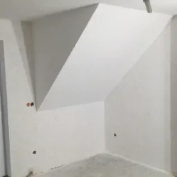 Surowe, białe wnętrze poddasza z widocznymi otworami na gniazdka elektryczne i wiszącą żarówką, przygotowane do dalszych prac wykończeniowych.