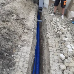 Wykop z ułożonymi niebieskimi rurami karbowanymi, prawdopodobnie peszlami, obok kostki brukowej i sterty ziemi, widoczny fragment instalacji przy budynku oraz osoba w roboczym ubraniu.