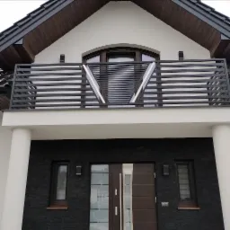 Nowoczesna, spawana balustrada balkonowa z poziomych profili, z geometrycznym wzorem w kształcie litery V, na tle elewacji domu z ciemnej cegły i białych kolumn.