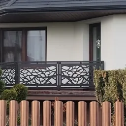 Balkon z dekoracyjną, ażurową balustradą w motywie liści, widoczny zza ogrodzenia z metalowych sztachet. Uwagę przykuwa nowoczesny design i dopasowanie do architektury domu.