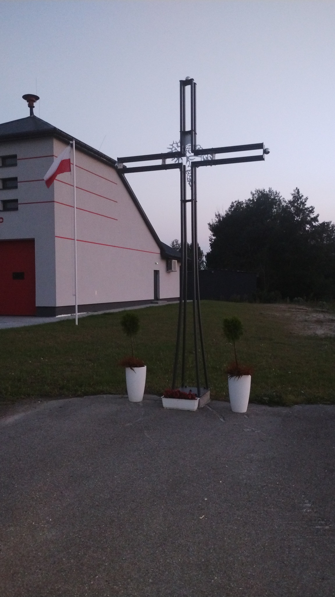 Metalowy krzyż z ozdobnymi elementami przed budynkiem straży pożarnej z flagą Polski, otoczony zielenią i donicami z kwiatami.