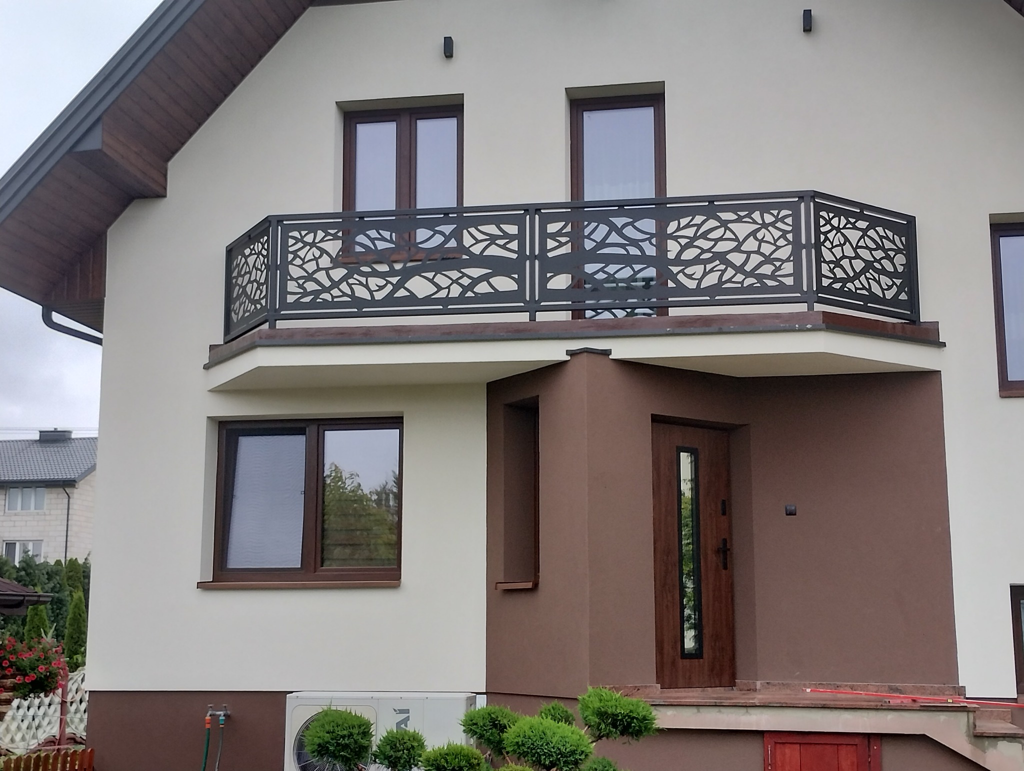 Elegancka, spawana balustrada balkonowa z motywem roślinnym, zamontowana na balkonie domu jednorodzinnego o jasnej elewacji, podkreślona brązowymi ramami okien i drzwiami.