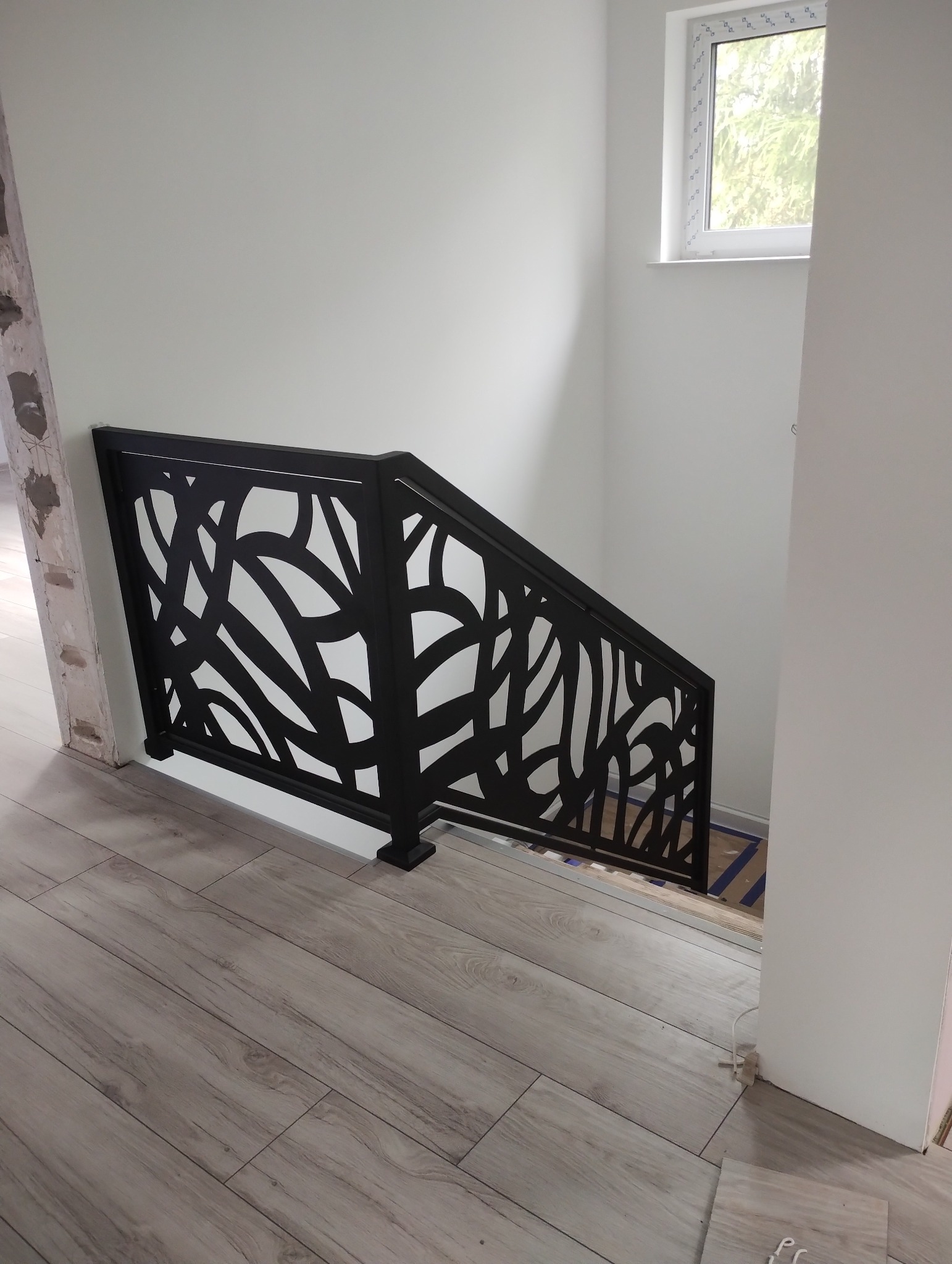 Nowoczesna, czarna, metalowa balustrada schodowa z ażurowym wzorem liści na tle białych ścian i drewnopodobnej podłogi.