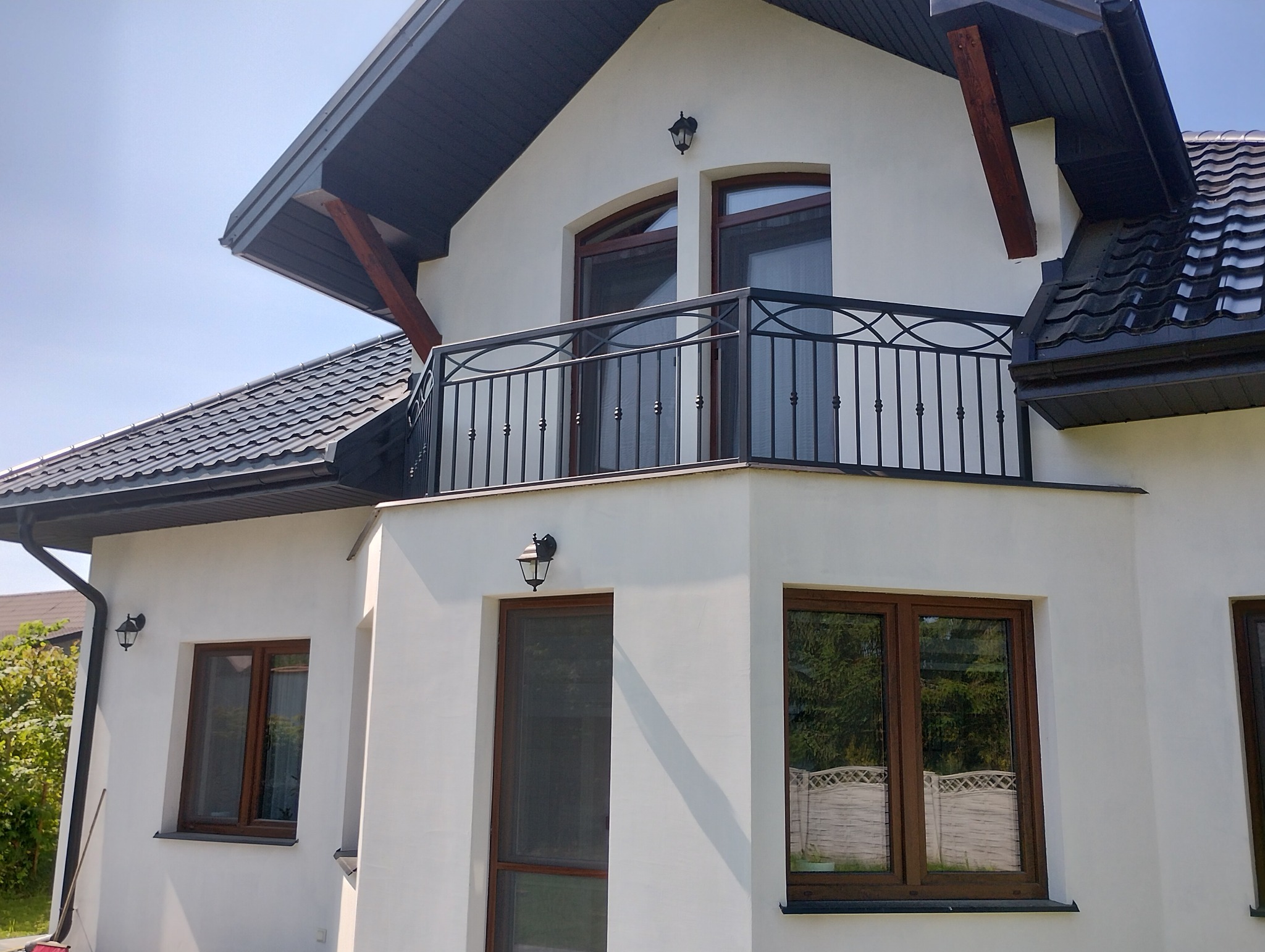Dwukondygnacyjny dom z białym tynkiem i czarną, kutą balustradą balkonową o dekoracyjnym wzorze. Widoczne okna z brązowymi ramami i dachówka w kolorze antracytu.