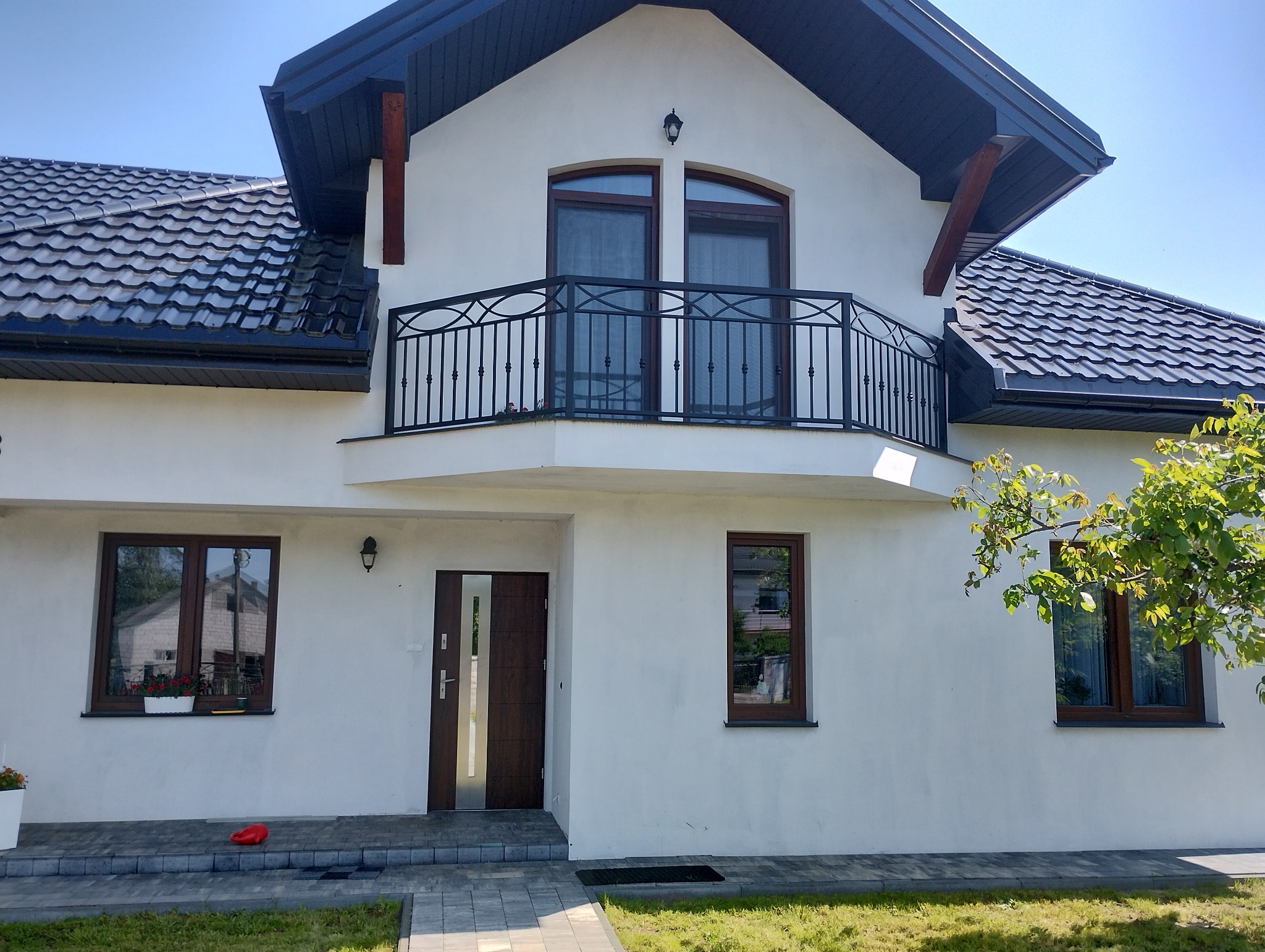 Biały dom z ciemną dachówką i balkonem z kutą balustradą. Widoczne okna z brązowymi ramami i drzwi wejściowe. Część domu otoczona zielenią.