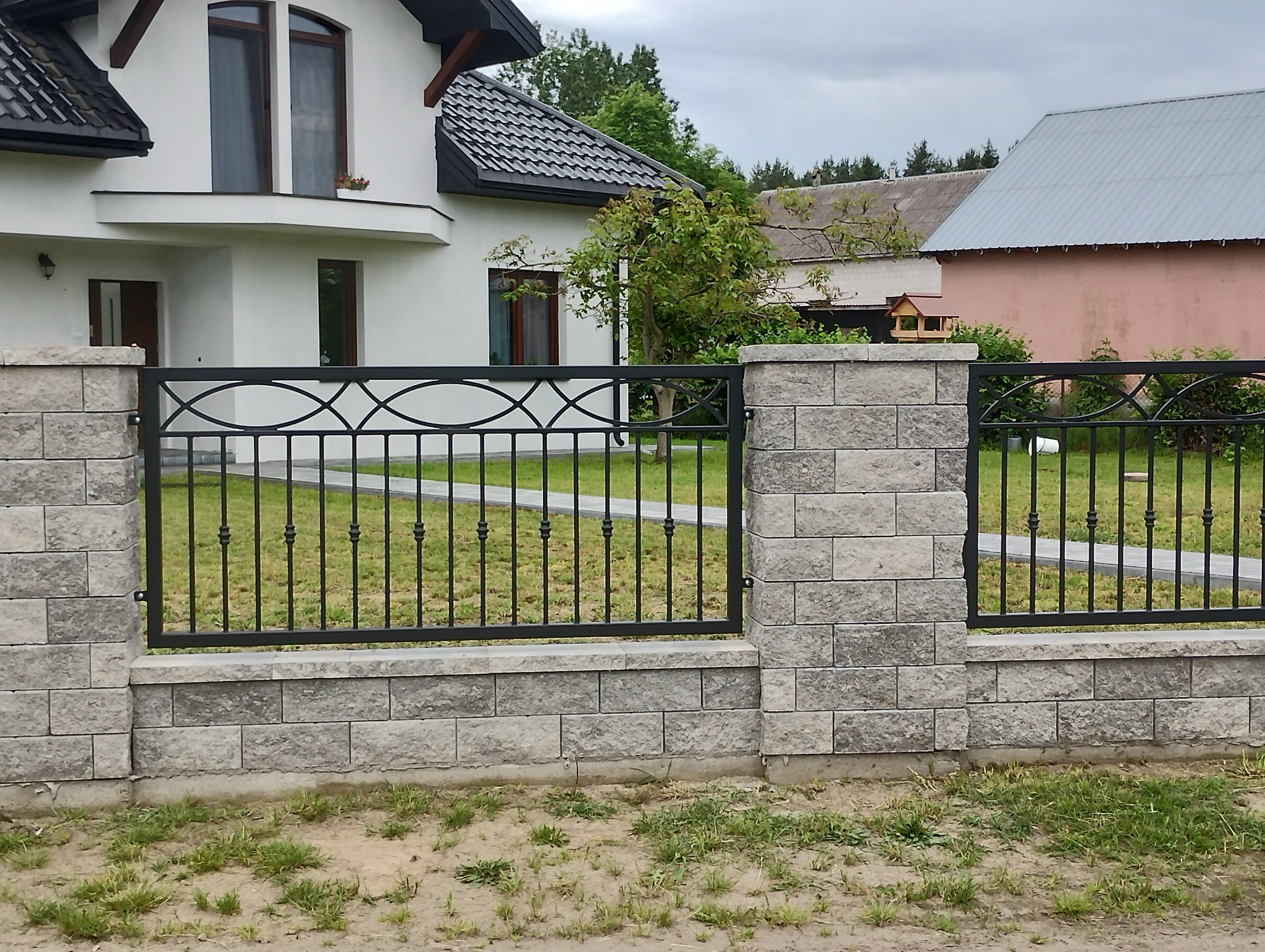 Nowoczesne ogrodzenie z metaloplastyki i kamiennych słupków, na tle domu z czarną dachówką. Precyzyjne wykonanie, estetyczne połączenie materiałów.
