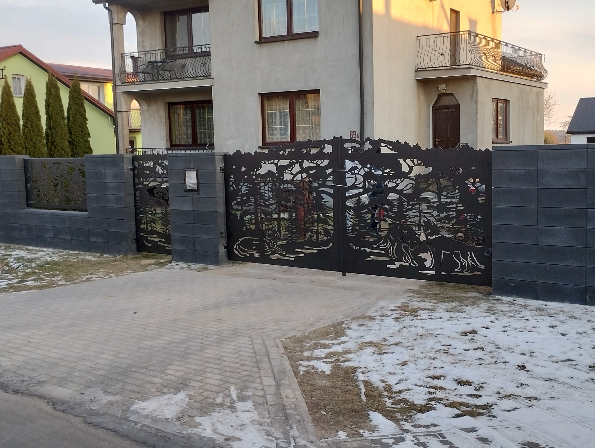 Metalowa brama z wycinanym laserowo motywem leśnym i wilkami, zamontowana przy nowoczesnym domu. Elementy spawane, szara kostka brukowa i fragmenty śniegu na trawniku.