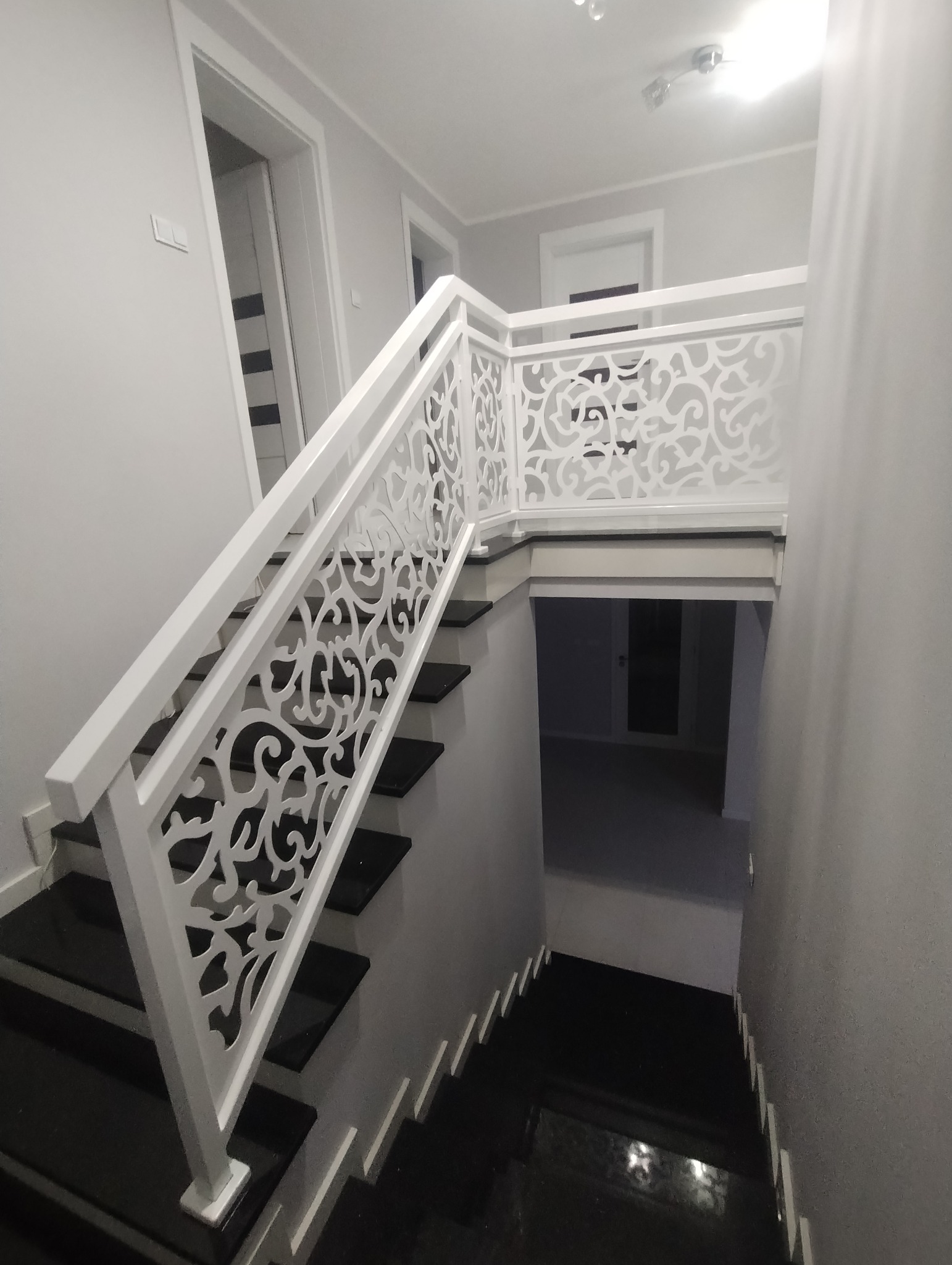 Nowoczesne, czarno-białe schody z ozdobną, ażurową balustradą w jasnym wnętrzu. Widok z góry na dół, z widocznym fragmentem podłogi i oświetleniem.