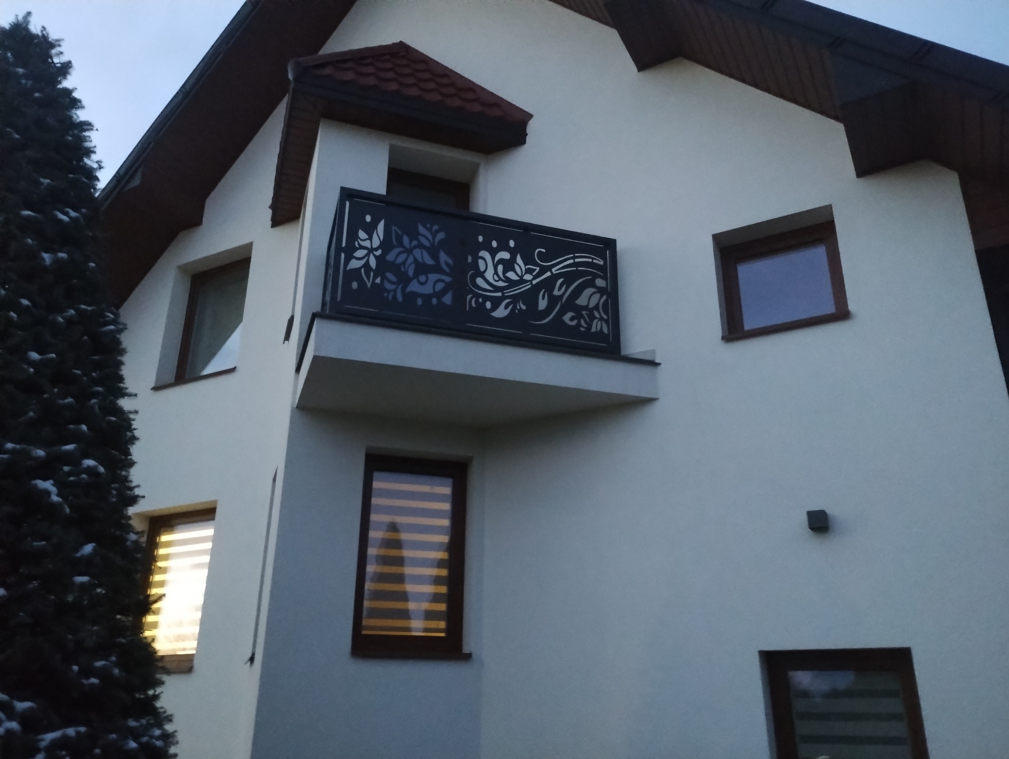 Biały dom z balkonem, którego ozdobna, metalowa balustrada z motywami roślinnymi została wykonana na zamówienie. Widok z dołu, w tle zielone drzewo.