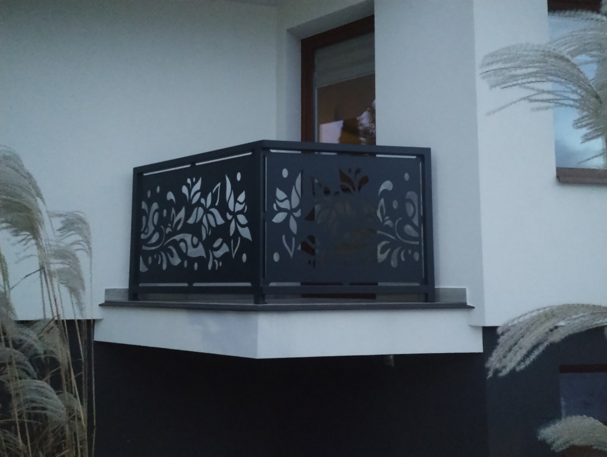 Metalowa balustrada balkonowa w kolorze grafitowym z laserowo wycinanymi motywami roślinnymi na tle białej elewacji budynku i dekoracyjnych traw.