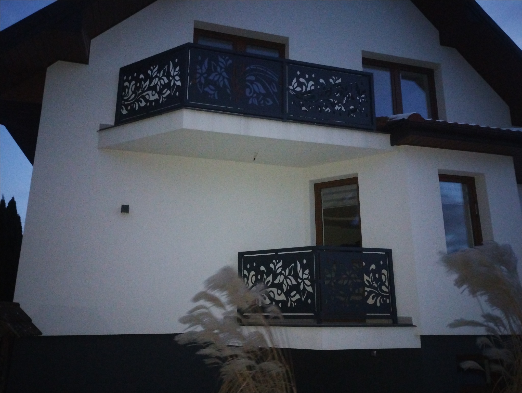 Eleganckie, metalowe balustrady balkonowe z wycinanym laserowo motywem roślinnym, zamontowane na białym budynku. Widoczne dwa balkony.